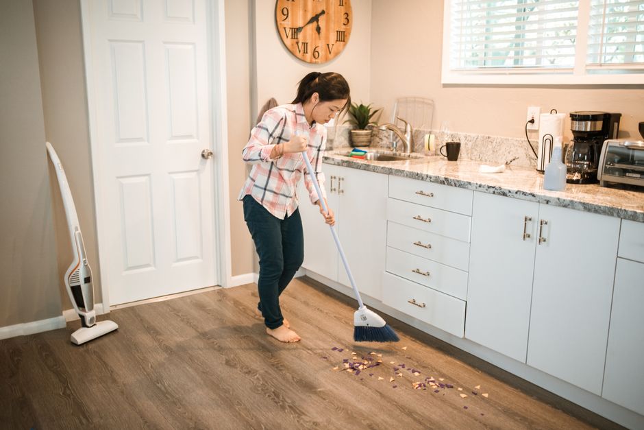 Asian woman sweeping kitchen floor with a broom, performing house chores in a modern kitchen.