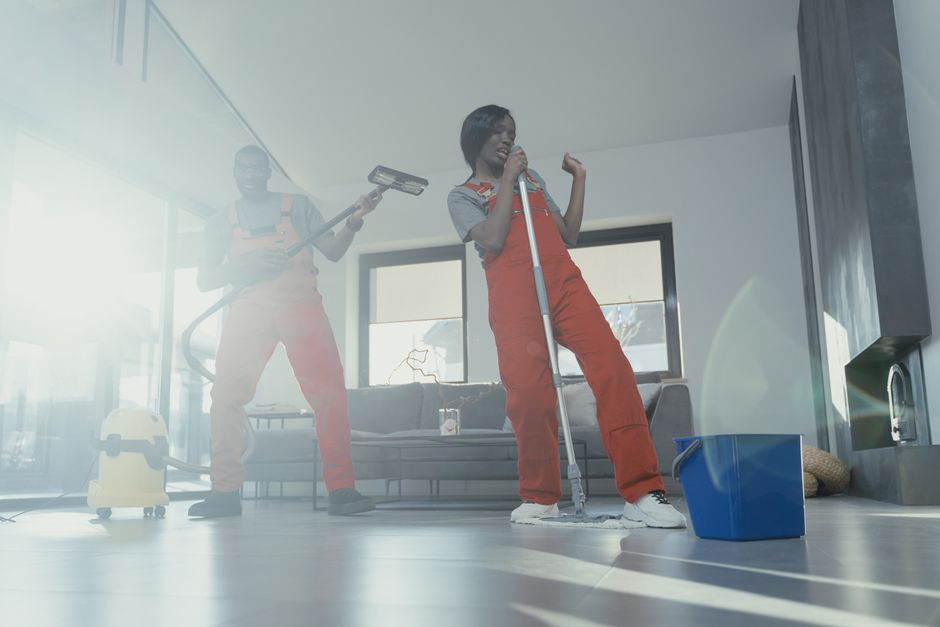 Two adults joyfully clean a modern home interior using a vacuum and mop, creating a lively and clean atmosphere.